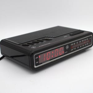 May include: A black GE digital clock radio with a red LED display showing 10:00. The device features a top-mounted speaker grille, control buttons, and a front-facing analog radio tuner dial with FM and AM frequency scales and the GE logo.