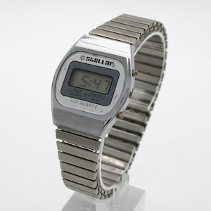 May include: A silver-toned digital wristwatch with a metallic expansion band. The watch face features the text Stellar, LCD Quartz, and a digital display showing 5:47. The screen includes labels for hour, min/sec, month, and date. Two small buttons are on the side.