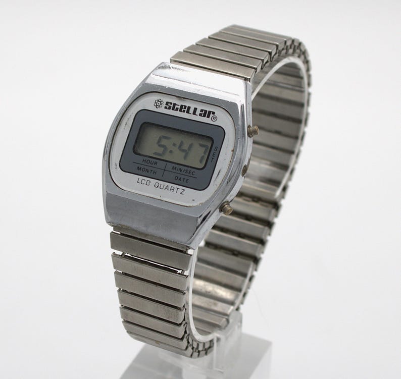 May include: A silver-toned digital wristwatch with a metallic expansion band. The watch face features the text Stellar, LCD Quartz, and a digital display showing 5:47. The screen includes labels for hour, min/sec, month, and date. Two small buttons are on the side.