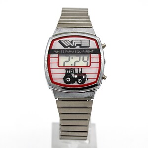 Peut inclure: Une montre num&eacute;rique vintage avec un bracelet en m&eacute;tal argent&eacute;. Le cadran de la montre affiche le texte "WFE WHITE FARM EQUIPMENT" au-dessus d'un affichage num&eacute;rique de l'heure. Un graphique de tracteur noir est en dessous, sur fond rouge et blanc.
