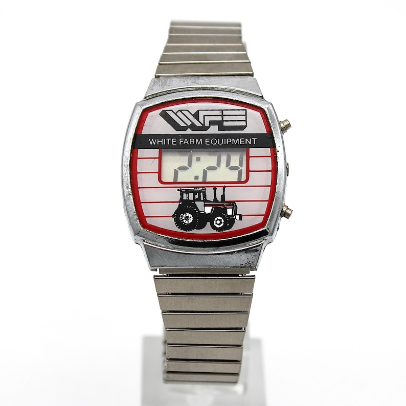 Peut inclure: Une montre num&eacute;rique vintage avec un bracelet en m&eacute;tal argent&eacute;. Le cadran de la montre affiche le texte "WFE WHITE FARM EQUIPMENT" au-dessus d'un affichage num&eacute;rique de l'heure. Un graphique de tracteur noir est en dessous, sur fond rouge et blanc.