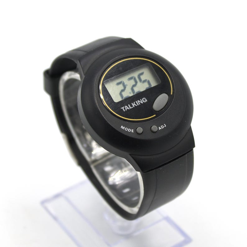 May include: Black digital talking watch with a round face and a black strap. The watch face displays the time and the word "TALKING". It has three buttons labelled "MODE" and "ADJ". The watch is displayed on a clear plastic stand.