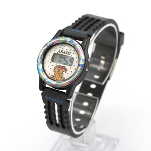 May include: Black digital watch with a black strap. The watch face has a colorful iridescent bezel, the brand name "ASAHI", and a cartoon bear. The strap has the word "SPORT" in blue.