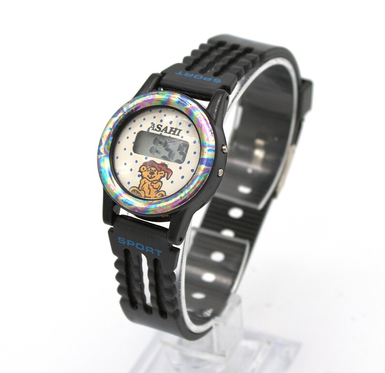 May include: Black digital watch with a black strap. The watch face has a colorful iridescent bezel, the brand name "ASAHI", and a cartoon bear. The strap has the word "SPORT" in blue.