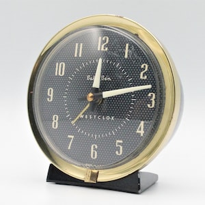 May include: A vintage "Westclox" "Ball Ben" alarm clock with a black face and gold-tone bezel. The clock has white hands and numerals, and sits on a black base. The clock face has a textured pattern.