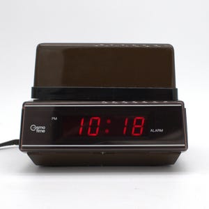 May include: A brown Cosmo Time digital alarm clock with a dark brown plastic casing and a black front panel. The display shows the time 10:18 in bright red LED digits, with a PM indicator on the left and an ALARM label on the right side.
