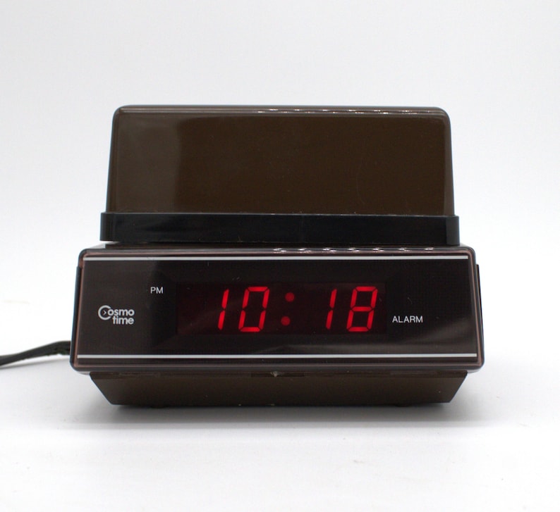 May include: A brown Cosmo Time digital alarm clock with a dark brown plastic casing and a black front panel. The display shows the time 10:18 in bright red LED digits, with a PM indicator on the left and an ALARM label on the right side.