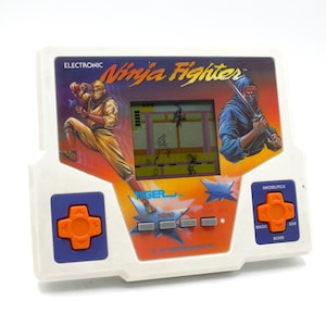 May include: A vintage Tiger Electronics "Ninja Fighter" handheld electronic game. The white plastic game features a small screen displaying a ninja fighting game, with orange and blue control buttons. The game's graphics include ninja illustrations and the text "Ninja Fighter".