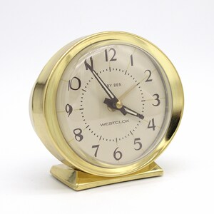 May include: A round gold-tone Westclox Baby Ben alarm clock with a cream-colored face, black Arabic numerals, and black hands. The clock features a clear protective cover and a rectangular gold-tone base, displaying the brand name Westclox.