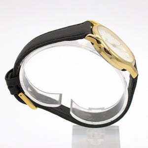 May include: A gold-toned wristwatch with a white face and black leather band. The watch has a classic design with a round face and a simple, elegant aesthetic. The band is secured with a gold-colored buckle.