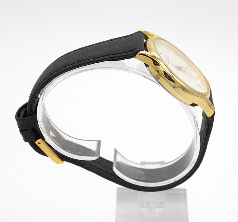 May include: A gold-toned wristwatch with a white face and black leather band. The watch has a classic design with a round face and a simple, elegant aesthetic. The band is secured with a gold-colored buckle.