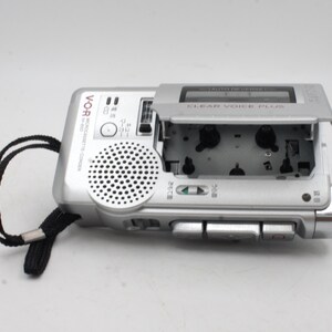 May include: A silver cassette recorder with a black strap. The cassette door is open, revealing the cassette tape inside. The recorder has a speaker on the front and several buttons on the side. The text "CLEAR VOICE PLUS" is printed on the top of the recorder.