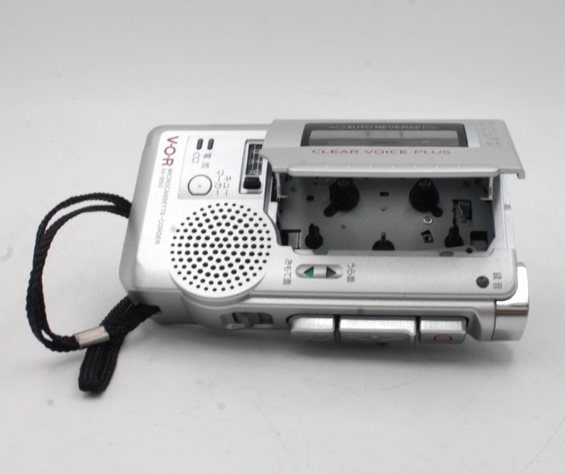 May include: A silver cassette recorder with a black strap. The cassette door is open, revealing the cassette tape inside. The recorder has a speaker on the front and several buttons on the side. The text "CLEAR VOICE PLUS" is printed on the top of the recorder.