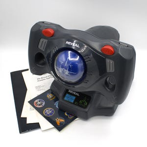 May include: A black and gray Reveal Flight Simulator toy with red buttons and a blue globe. The toy has the word "Reveal" printed on it. Included are instruction sheets and sticker sheets with various emblems.