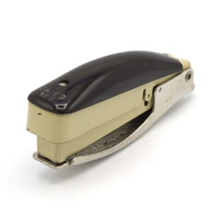 May include: A vintage stapler with a black top, beige body, and silver metal base. The stapler has the text "BATES" and "BATES 80" printed on it. The design is a classic office supply.