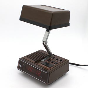 May include: A brown Cosmo Time digital alarm clock with an integrated adjustable lamp. The base features a red digital display showing 10:12, a speaker grille, and black control buttons. A metallic hinged arm connects the base to the rectangular lamp.