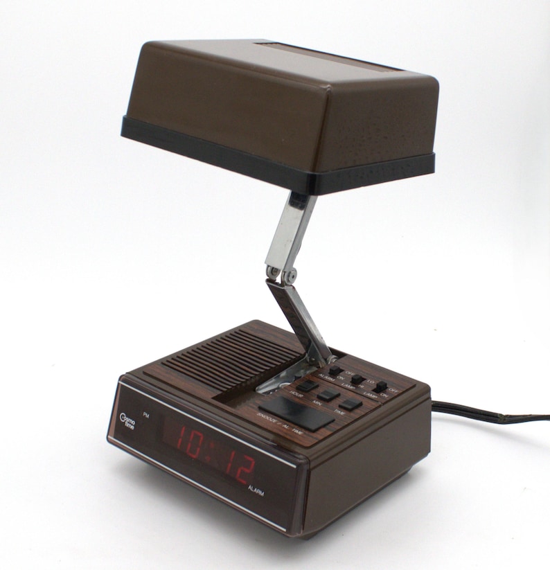 May include: A brown Cosmo Time digital alarm clock with an integrated adjustable lamp. The base features a red digital display showing 10:12, a speaker grille, and black control buttons. A metallic hinged arm connects the base to the rectangular lamp.