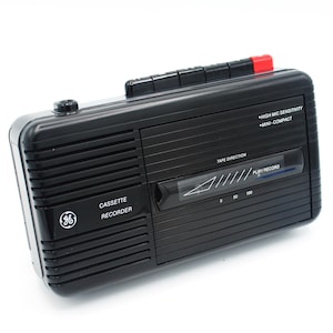 May include: A black cassette recorder with a silver GE logo and the text "CASSETTE RECORDER" on the front. The recorder has a tape direction indicator and a play/record button. The text "HIGH MIC SENSITIVITY" and "MINI-COMPACT" are printed on the side.