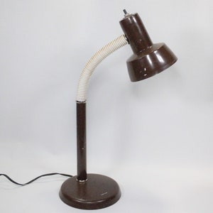 May include: A brown desk lamp with a flexible, white, ribbed neck. The lamp has a round base and a conical shade. The lamp is plugged into a black electrical cord. The lamp is approximately 15 inches tall.