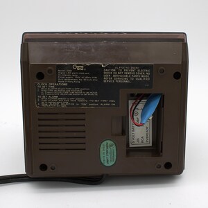 May include: The back of a brown Cosmo Time digital LED alarm clock, model 5501, featuring a black instructional label for clock operations, a 9-volt battery compartment with a blue connector, a power cord, and a green UL portable lamp sticker.