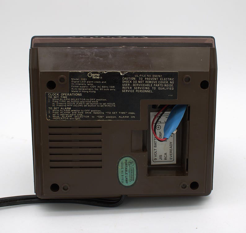May include: The back of a brown Cosmo Time digital LED alarm clock, model 5501, featuring a black instructional label for clock operations, a 9-volt battery compartment with a blue connector, a power cord, and a green UL portable lamp sticker.