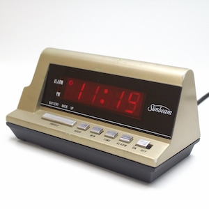 May include: A gold-colored digital alarm clock with a black face and red LED display showing the time 11:19. The clock has a Sunbeam logo and buttons for snooze, hour, minute, time, alarm, on, and off.