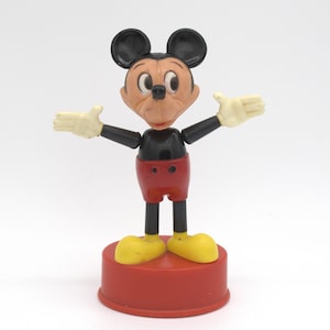 May include: A vintage Mickey Mouse figurine standing on a circular red base. The character has a peach-colored face, large black ears, a black torso, red shorts with two black buttons, and yellow shoes. The arms are outstretched with white gloves.