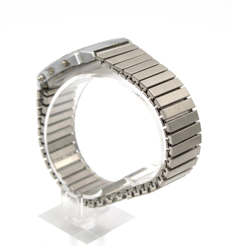 Peut inclure: Une montre-bracelet argent&eacute;e avec un cadran rectangulaire et un bracelet en m&eacute;tal flexible. Le bracelet est compos&eacute; de nombreux maillons rectangulaires. La montre est pr&eacute;sent&eacute;e sur un support transparent sur fond blanc.