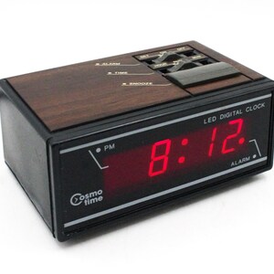 May include: A black and wood-toned digital alarm clock with a red LED display showing the time 8:12 PM. The clock has a Cosmo Time logo and the words "LED Digital Clock" and "Alarm" printed on it.