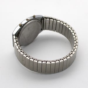 May include: A silver-toned digital wristwatch featuring a round case with three small side buttons and a flexible, expandable metal link bracelet. The watch is shown from the back, displaying a plain, circular stainless steel case back.