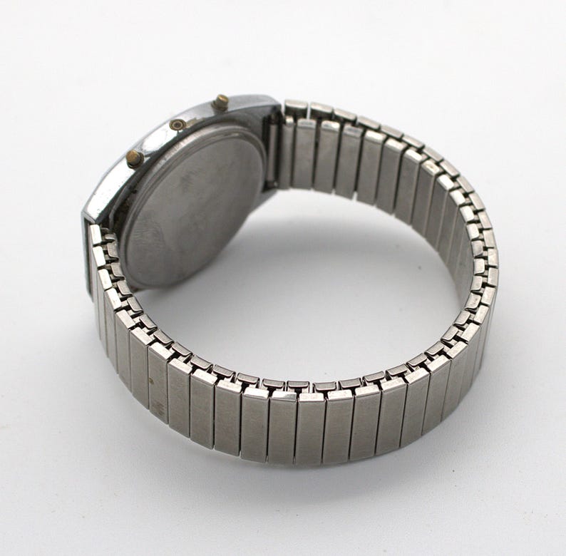 May include: A silver-toned digital wristwatch featuring a round case with three small side buttons and a flexible, expandable metal link bracelet. The watch is shown from the back, displaying a plain, circular stainless steel case back.