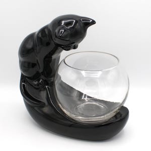 May include: A black ceramic cat-shaped planter with a clear glass bowl. The cat is glossy and perched, looking down at the bowl. The base is a matching black, oval shape. A decorative item for plants or small items.
