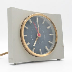 May include: A gray and gold alarm clock with a blue face and white hour and minute hands. The clock has a simple design and is a great addition to any bedside table.