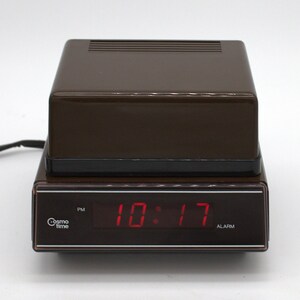 May include: A brown rectangular digital alarm clock with a black base and a dark display panel. The front features the text Cosmo time, a PM indicator, and the red digital time display 10:17. The top has a ribbed surface and a power cord extends out.