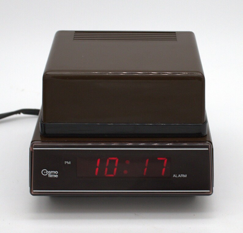 May include: A brown rectangular digital alarm clock with a black base and a dark display panel. The front features the text Cosmo time, a PM indicator, and the red digital time display 10:17. The top has a ribbed surface and a power cord extends out.
