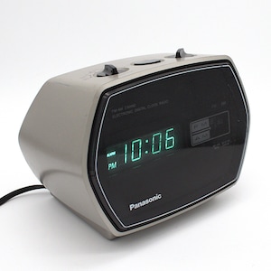 May include: A vintage Panasonic FM-AM electronic digital clock radio. The clock has a gray body with a black front panel displaying the time in green digits. The time reads 10:06 PM. The clock has two top buttons and the brand name is visible.
