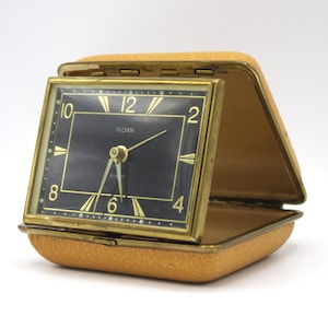 May include: A vintage gold-toned alarm clock with a black face and white numerals. The clock is in a yellow leather case and has the word "FLORN" printed on the face.