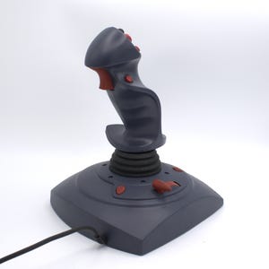 Puede incluir: Un joystick gris con detalles rojos, que consta de una base, un eje central con una bota de goma negra y un mango con botones. Un cable negro se extiende desde la base. El joystick est&aacute; dise&ntilde;ado para juegos o simulaci&oacute;n de vuelo.