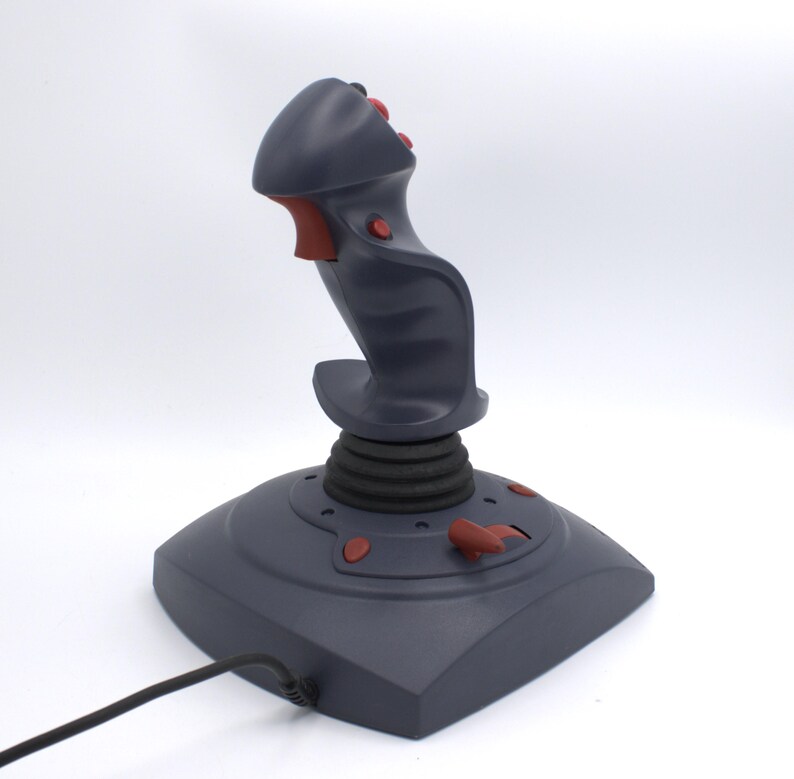 Puede incluir: Un joystick gris con detalles rojos, que consta de una base, un eje central con una bota de goma negra y un mango con botones. Un cable negro se extiende desde la base. El joystick est&aacute; dise&ntilde;ado para juegos o simulaci&oacute;n de vuelo.