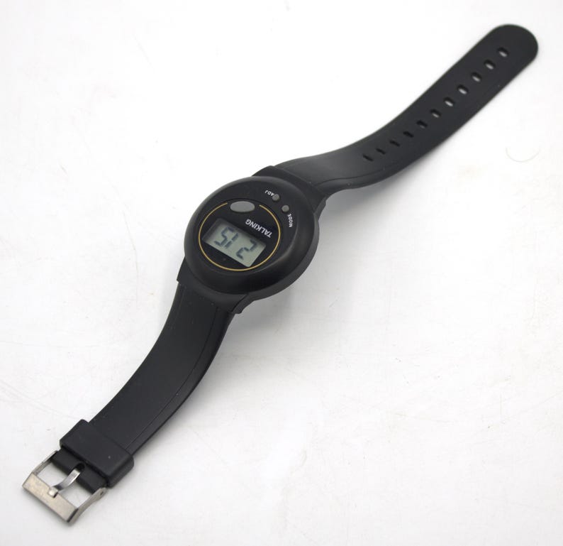May include: Black digital talking watch with a black strap and silver buckle. The watch face displays the time, and the word "TALKING" is printed on the face. The strap is made of a flexible material.