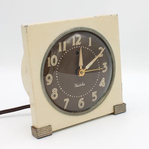 May include: A vintage white electric clock with a brown face and black hands. The clock has a square shape with rounded corners and a cord for plugging it in. The clock face has the word "Westelox" printed on it.