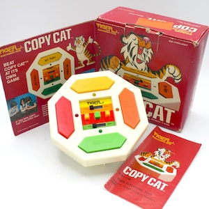 May include: A vintage Tiger Electronic Toys Copy Cat game with a white octagonal base and colorful buttons. The game features a digital display and a red, yellow, green, and orange button. The game is packaged in a red box with a cartoon tiger on the front.