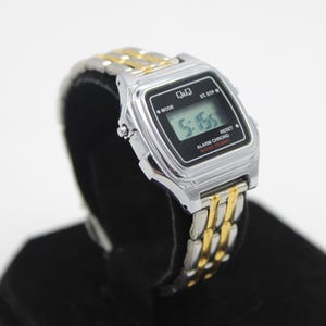 May include: A silver and gold two-tone digital wristwatch with a black face. The watch face displays the time as 5:16. The watch has a black band with a silver and gold two-tone buckle. The watch has the words "Alarm Chrono Water Resist" printed on the face.