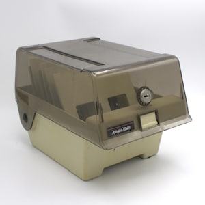 May include: A beige and brown plastic Media Mate card file box with a clear, tinted lid. The box has a lock and a label that reads "Media Mate". The box is designed to hold cards.