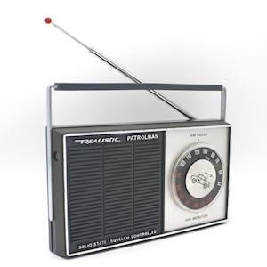 May include: A silver and black Realistic Patrolman AM radio with a telescopic antenna. The radio has a dial with a police car graphic and the text "AM Radio" and "VHF Monitor". The text "SOLID STATE SQUELCH CONTROLLED" is printed on the bottom of the radio.