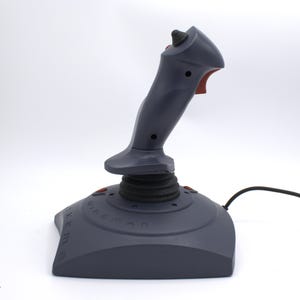 Puede incluir: Un joystick gris con gatillo y botones rojos. La base es de color gris oscuro con el texto "WingMan" y "Extreme" impreso. Un cable negro se extiende desde la base.
