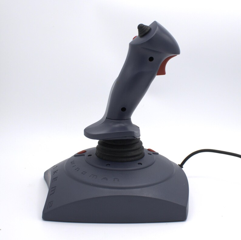 Puede incluir: Un joystick gris con gatillo y botones rojos. La base es de color gris oscuro con el texto "WingMan" y "Extreme" impreso. Un cable negro se extiende desde la base.