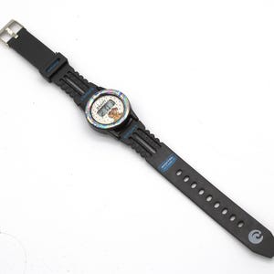 May include: Black digital watch with a round face featuring a cartoon character. The watch has a black strap with a silver buckle. The watch face has a rainbow-colored bezel and the word "SPORT" is printed on the strap.