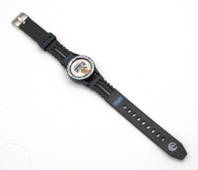 May include: Black digital watch with a round face featuring a cartoon character. The watch has a black strap with a silver buckle. The watch face has a rainbow-colored bezel and the word "SPORT" is printed on the strap.