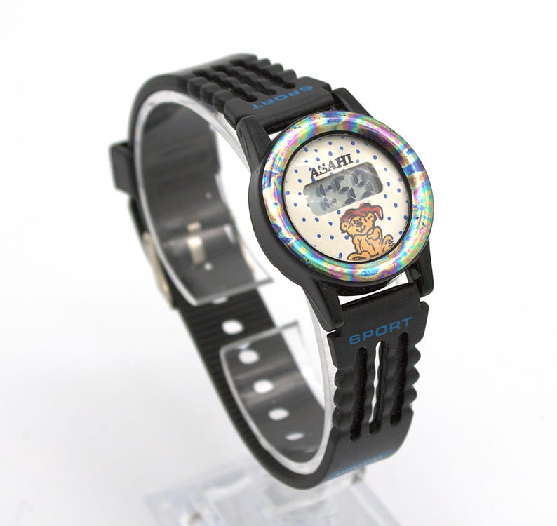 May include: Black digital watch with a black band. The watch face has a cartoon dog illustration and the word "ASAHI". The watch band has the word "SPORT" printed on it.
